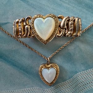 May include: A gold-toned jewelry set featuring a heart-shaped pendant necklace and a matching bracelet. Both pieces have a mother-of-pearl heart center, framed by a decorative gold border. The bracelet is a stretch style.