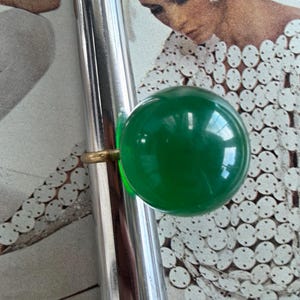 May include: A vintage ring featuring a large, round, bright green stone. The stone is set on a thin, gold-colored band. The ring is displayed on a silver metal object, with a vintage magazine in the background.