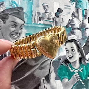 May include: A gold-toned vintage bracelet with a heart-shaped locket. The bracelet has a flexible, ribbed design. The locket features an embossed floral design. The background is a vintage illustration with figures in military uniforms and a young girl.