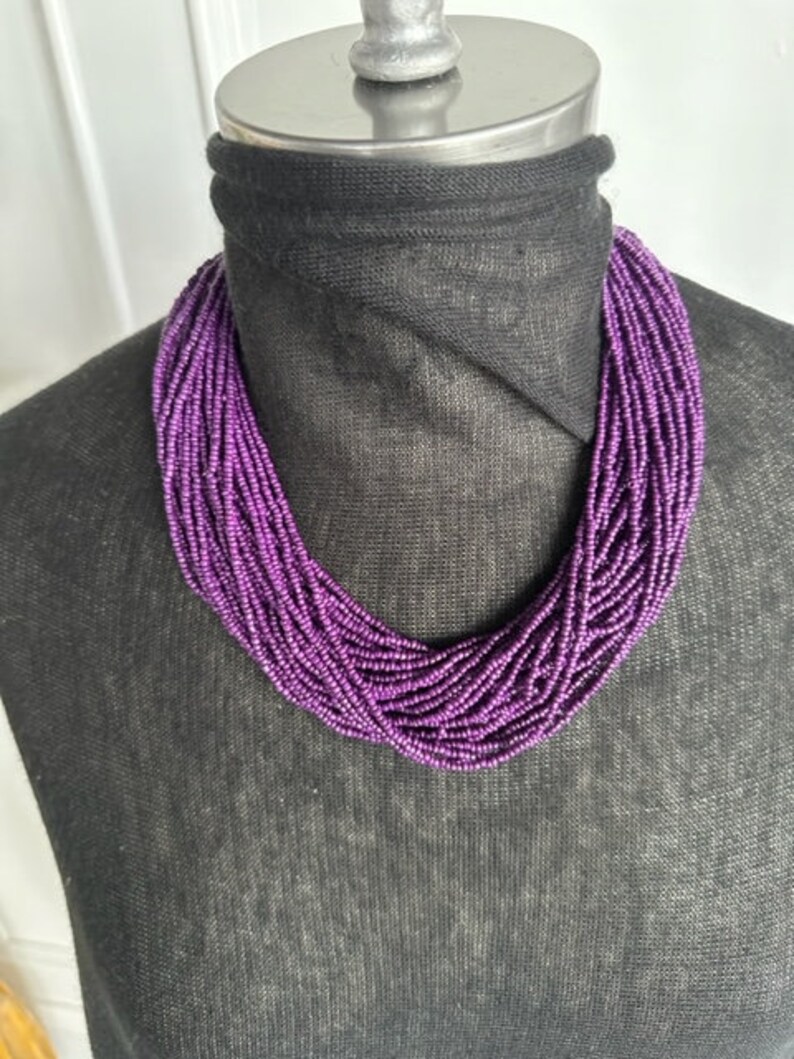May include: A purple beaded necklace with multiple strands of small beads. The necklace is displayed on a black mannequin.