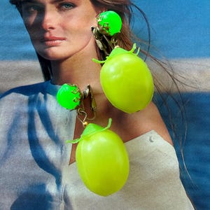 May include: A pair of clip-on earrings featuring bright neon green spherical beads set in gold-tone metal prongs, each dangling a large, smooth, oval-shaped neon green fruit-like charm with a small green leaf detail at the top.