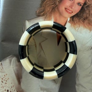 May include: A thick, circular bangle bracelet featuring a repeating checkerboard pattern of alternating black and off-white squares. The bracelet has a smooth, polished finish and a wide, chunky profile that creates a bold, high-contrast geometric design.