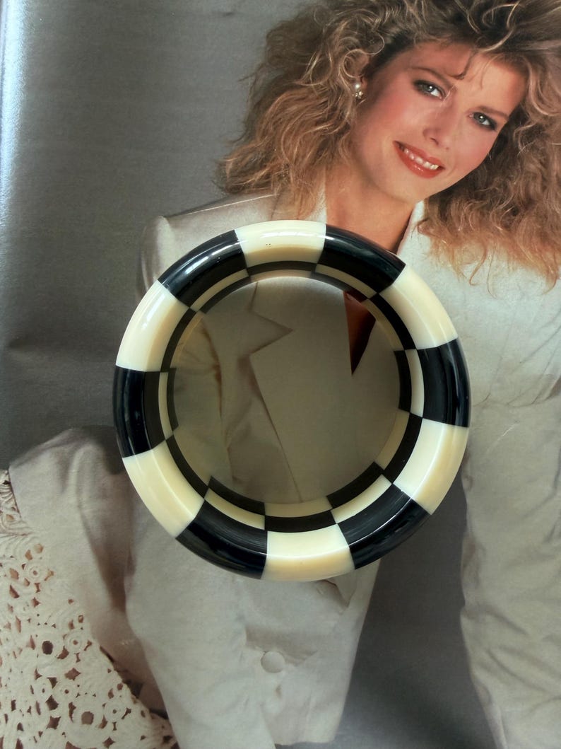 May include: A thick, circular bangle bracelet featuring a repeating checkerboard pattern of alternating black and off-white squares. The bracelet has a smooth, polished finish and a wide, chunky profile that creates a bold, high-contrast geometric design.