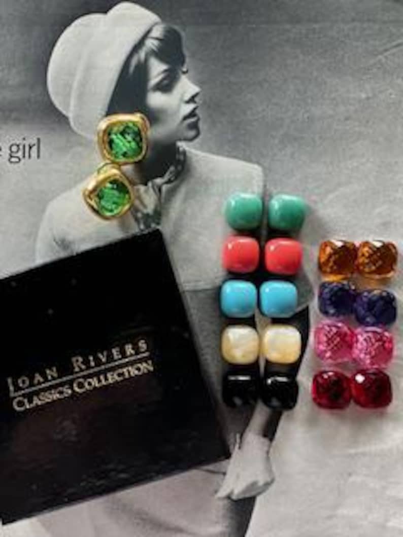 May include: A collection of Joan Rivers Classics Collection earrings. The earrings are square-shaped with gold-tone metal and colorful, faceted stones. The colors include green, red, blue, white, black, and various shades of pink and orange. A black box with gold lettering is also present.
