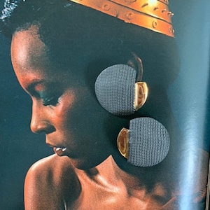 May include: A pair of large, round earrings with a woven fabric texture in a dark gray color. The earrings have a gold-toned metal accent on the side.