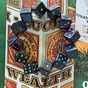 May include: A circular bracelet composed of twelve black dice, each featuring pips in various colors including blue, yellow, green, orange, and pink. The dice are arranged in a ring shape, showcasing a variety of colored dots on their surfaces.
