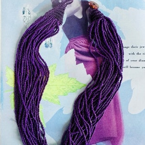 May include: A long purple beaded necklace with a tassel at the end. The necklace is made of small, round beads that are strung together in a long strand. The tassel is made of the same beads, but they are gathered together at the end of the strand.