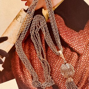 May include: A gold-toned necklace with multiple strands of delicate chain. The necklace features a decorative pendant with a spherical cage design and a tassel. The background includes a person wearing a red tweed jacket.