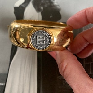 May include: Gold-tone bangle bracelet with a circular, black and silver logo that reads "Paris New York Givenchy".