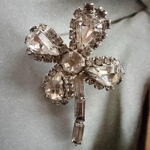 May include: A silver-toned brooch in a floral design, featuring clear, faceted crystals. The brooch has a central round crystal, with four larger crystals forming the petals. The stem is composed of rectangular crystals.