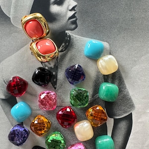 May include: A collection of colorful, square-shaped vintage earrings. The earrings feature a variety of colors, including red, pink, green, blue, and gold. Some earrings have a faceted design, while others are smooth. The background is a black and white photograph.