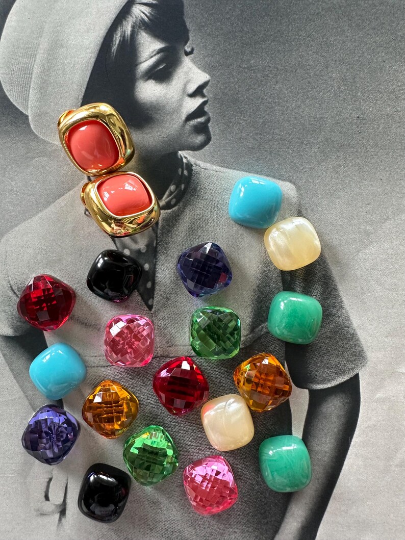 May include: A collection of colorful, square-shaped vintage earrings. The earrings feature a variety of colors, including red, pink, green, blue, and gold. Some earrings have a faceted design, while others are smooth. The background is a black and white photograph.