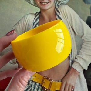 May include: A bright yellow, glossy, wide bangle bracelet. The bracelet is a solid, curved shape, reflecting light. The image is of a vintage style, with a person in the background wearing a striped hat and a yellow belt.