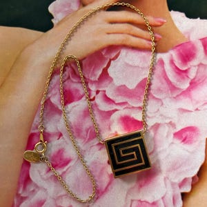 May include: A gold-toned necklace with a square pendant featuring a black and gold geometric design. The necklace is displayed against a backdrop of pink and white floral fabric, creating a vintage aesthetic.