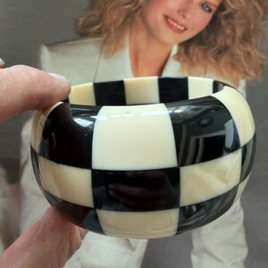 May include: A wide bangle bracelet featuring a high-gloss finish with a repeating black and off-white checkered pattern. The circular accessory has a smooth, rounded surface and a thick band construction that displays a bold, contrasting geometric design.