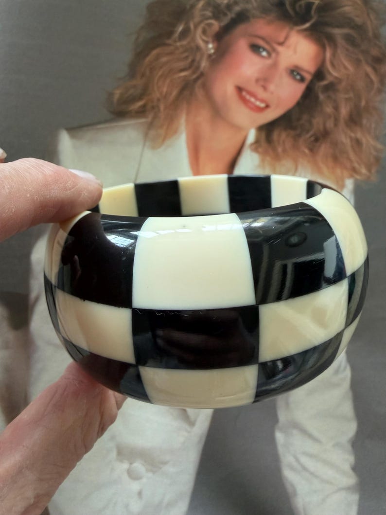 May include: A wide bangle bracelet featuring a high-gloss finish with a repeating black and off-white checkered pattern. The circular accessory has a smooth, rounded surface and a thick band construction that displays a bold, contrasting geometric design.