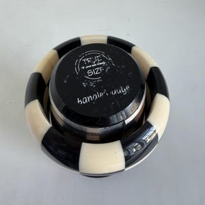 May include: A black and white checkered bangle gauge tool with a circular top featuring the text TRUE SIZE and bangle gauge. The tool displays a measurement of 6.67 inches and 169.4 mm on its side, used for determining the size of a circular bracelet.