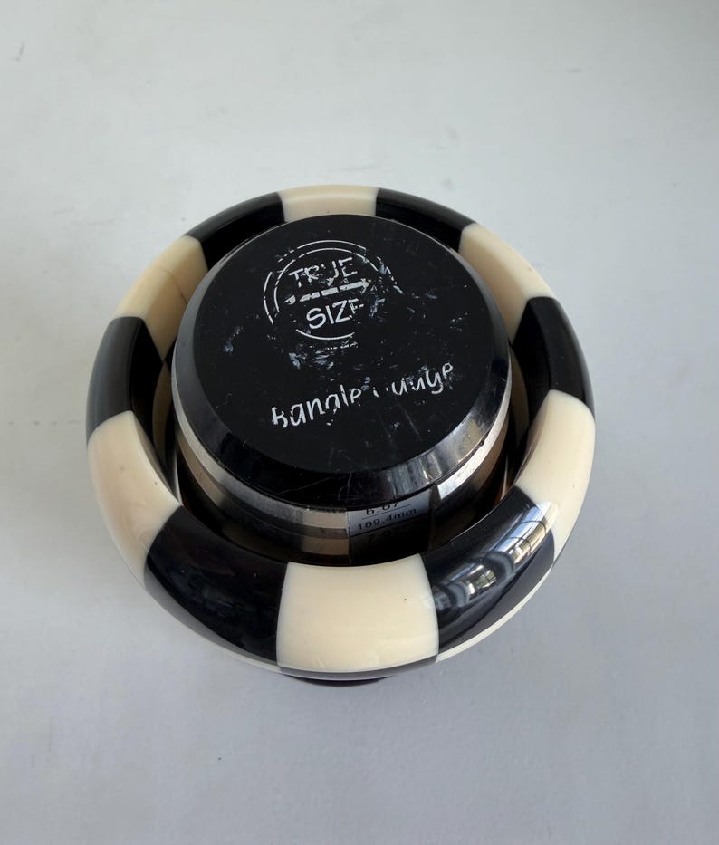 May include: A black and white checkered bangle gauge tool with a circular top featuring the text TRUE SIZE and bangle gauge. The tool displays a measurement of 6.67 inches and 169.4 mm on its side, used for determining the size of a circular bracelet.