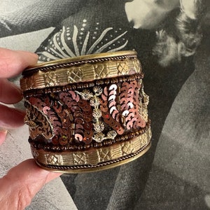 May include: A gold metal cuff bracelet with a beaded and sequined design. The sequins are in shades of brown, pink, and gold.