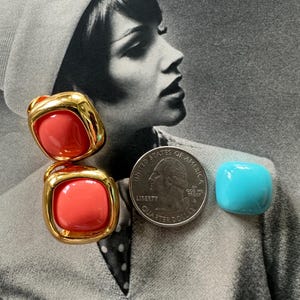 May include: Two coral-colored, square-shaped earrings with gold-tone metal frames. A quarter coin and a small, turquoise-colored square are also in the image. The background is a black and white photograph of a woman.