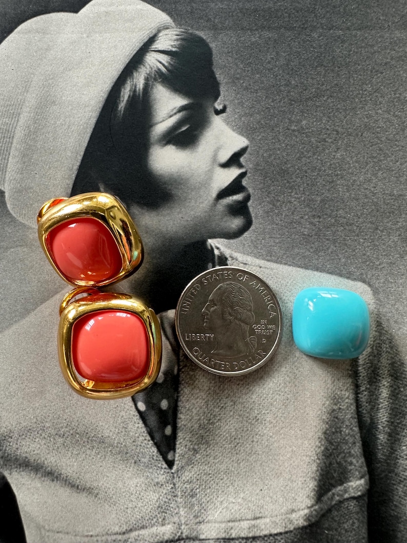 May include: Two coral-colored, square-shaped earrings with gold-tone metal frames. A quarter coin and a small, turquoise-colored square are also in the image. The background is a black and white photograph of a woman.
