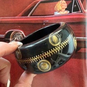May include: A black bangle bracelet with gold-colored zipper and circular accents. The bracelet is held in front of a red car with a woman inside. The bracelet is a bold fashion accessory.