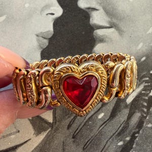 May include: Gold-toned stretch bracelet featuring a large, heart-shaped red gemstone. The heart is framed by a decorative gold border, and the bracelet has an ornate, textured design. The background is a black and white image.