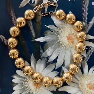May include: A gold-colored beaded necklace with a chain clasp. The necklace features large, round beads that reflect light. The necklace is displayed on a dark blue background with white floral accents.