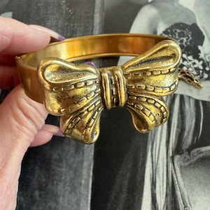 May include: A gold-toned metal bangle bracelet featuring a prominent bow design. The bow has detailed texturing, and the bracelet has a hinged closure. The overall aesthetic is vintage-inspired, with a slightly aged appearance.