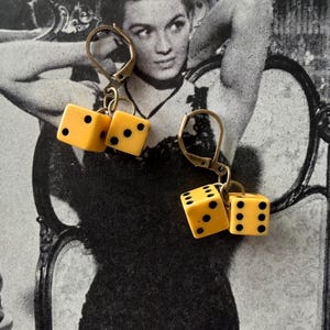 May include: A pair of vintage-style earrings featuring small, yellow dice with black dots. The dice are suspended from gold-colored lever-back closures. The background is a black and white image of a woman.