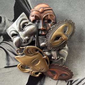 May include: A collection of antique metal masks in various styles and colors. The masks include a silver comedy mask, a copper-toned mask with a face, and several decorative masks with gold and bronze finishes. The masks are arranged on a black and white background.