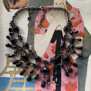 May include: A multi-layered statement necklace featuring dark gunmetal-toned chain links and numerous faceted black oval and teardrop-shaped beads. The design includes a central cluster of stones with dangling pendants and a lobster claw clasp closure.