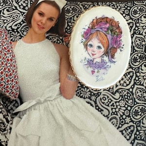 May include: A white oval ceramic plate with a gold rim features a hand-painted illustration of a girl with purple flowers in her hair. The girl has blue eyes, red hair, and is wearing a purple dress. The plate has the word "Leslie" written on the bottom.