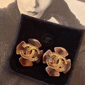 Puede incluir: Un par de pendientes con forma de flor con el logotipo CC entrelazado en tono dorado en el centro. Los p&eacute;talos son de color marr&oacute;n y beige transl&uacute;cido. Los pendientes se muestran sobre una superficie de terciopelo negro.