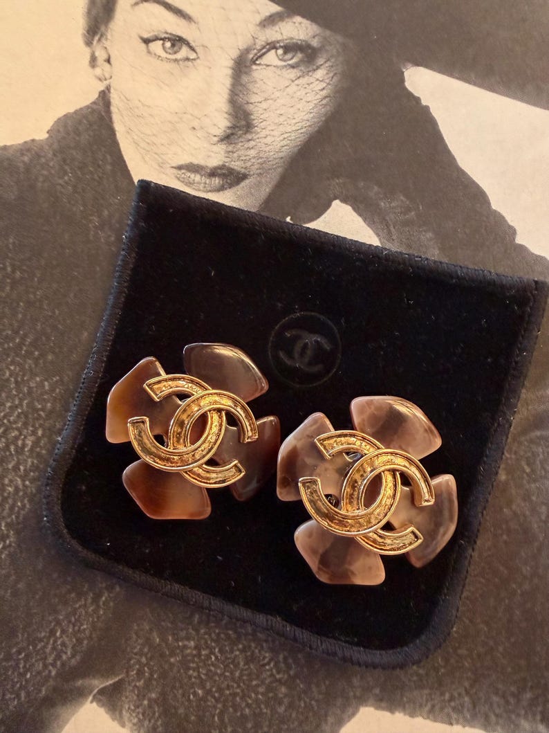 Puede incluir: Un par de pendientes con forma de flor con el logotipo CC entrelazado en tono dorado en el centro. Los p&eacute;talos son de color marr&oacute;n y beige transl&uacute;cido. Los pendientes se muestran sobre una superficie de terciopelo negro.