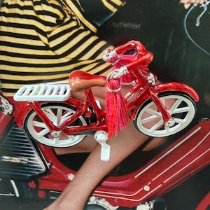 May include: A miniature red motorcycle with white wheels and a brown seat. The motorcycle has a decorative tassel and is set against a background with a person wearing a black and yellow striped top and yellow flats.