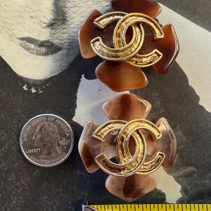 Puede incluir: Dos pendientes marrones y dorados con forma de flor. Cada pendiente presenta un logotipo doble C entrelazado dorado en el centro. Los pendientes miden aproximadamente 3,8 cm de largo. Una moneda est&aacute; al lado.