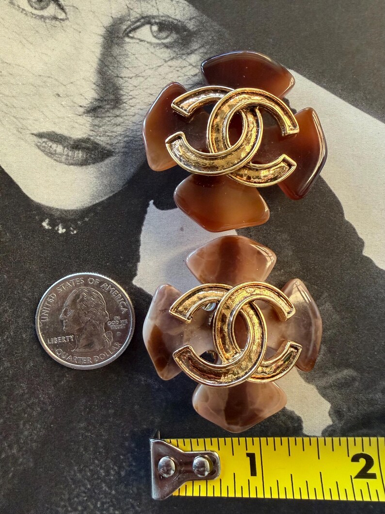 Puede incluir: Dos pendientes marrones y dorados con forma de flor. Cada pendiente presenta un logotipo doble C entrelazado dorado en el centro. Los pendientes miden aproximadamente 3,8 cm de largo. Una moneda est&aacute; al lado.