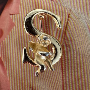 May include: Gold-toned metal brooch in the shape of the letter "S" with a small cherub figure. The cherub holds a small object. The brooch is pinned to a pink and yellow striped fabric.