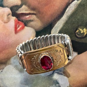 May include: Vintage gold-tone bracelet with a central oval-cut red gemstone. The bracelet features an engraved floral design on the gold plate and a silver-tone, expandable band. A classic piece of jewelry.
