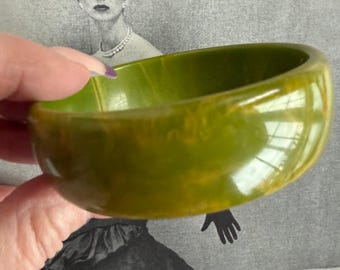 May include: A vintage olive green bangle bracelet. The bracelet is a solid, rounded shape with a smooth, glossy finish. The color has a marbled effect with hints of yellow. The background features a vintage black and white image of a woman.