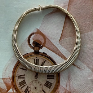 May include: A gold-colored, ribbed collar necklace with a clasp. The necklace is circular and sits atop a vintage pocket watch illustration with a cream-colored face and Roman numerals. The background includes a sheer, peach-colored fabric.