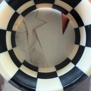 May include: A circular bangle bracelet featuring a repeating black and white checkered pattern. The accessory has a smooth, polished finish and a wide band design that creates a bold, high-contrast geometric look around the entire circumference.