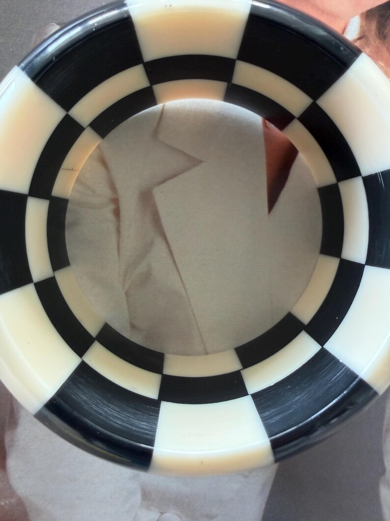 May include: A circular bangle bracelet featuring a repeating black and white checkered pattern. The accessory has a smooth, polished finish and a wide band design that creates a bold, high-contrast geometric look around the entire circumference.