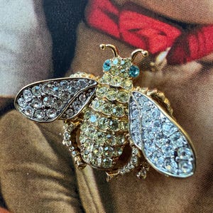 May include: A gold-toned bee brooch encrusted with clear and yellow rhinestones. The wings are detailed with clear rhinestones, and the body features yellow rhinestones. The bee has small blue rhinestone eyes and delicate antennae.