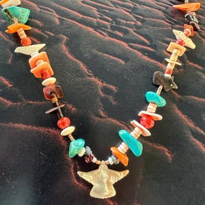 May include: A multi-strand necklace featuring a central carved shell bird pendant. The necklace is composed of various beads including turquoise, orange coral, white shell, and dark stone accents, interspersed with small cylindrical spacers.