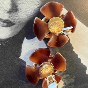 Puede incluir: Un par de pendientes vintage de clip. Cada pendiente presenta un dise&ntilde;o floral con cinco p&eacute;talos marrones. El centro de cada pendiente tiene un disco de metal dorado con texto y un mecanismo de clip.