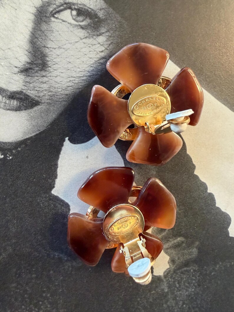 Puede incluir: Un par de pendientes vintage de clip. Cada pendiente presenta un dise&ntilde;o floral con cinco p&eacute;talos marrones. El centro de cada pendiente tiene un disco de metal dorado con texto y un mecanismo de clip.