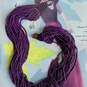 May include: A long, multi-strand necklace made of small, purple beads. The necklace is draped over a magazine with a woman on the cover.