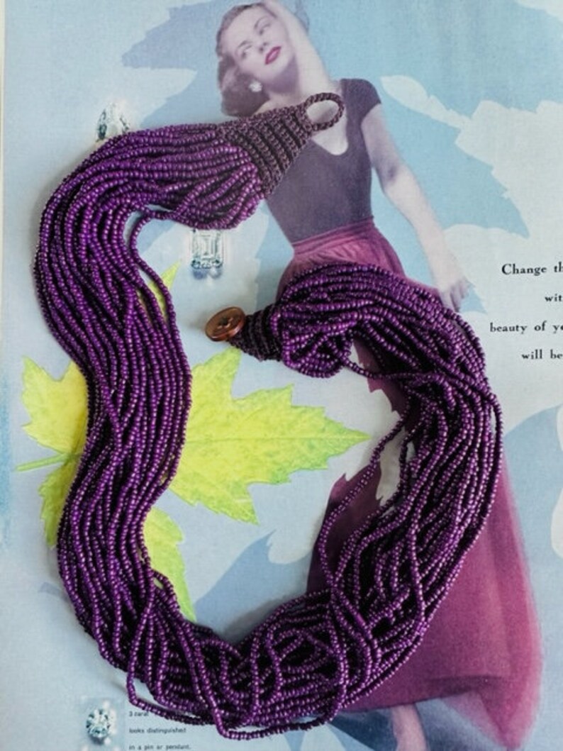 May include: A long, multi-strand necklace made of small, purple beads. The necklace is draped over a magazine with a woman on the cover.
